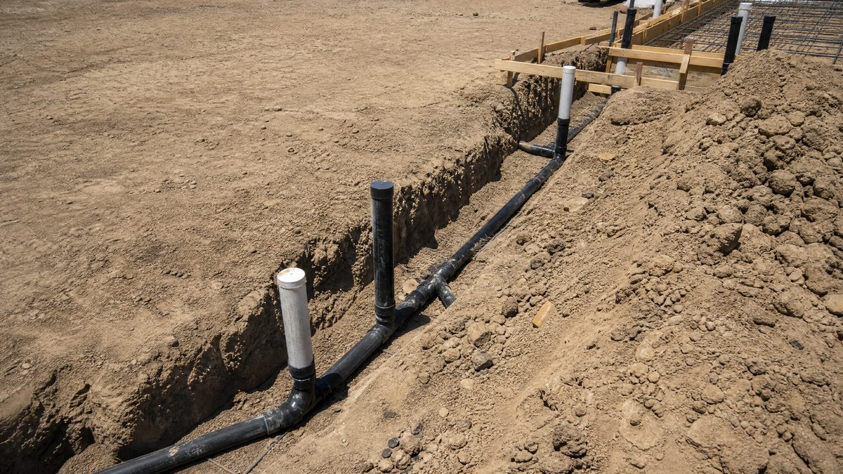 Underground plumbing rough-in trench with ABS drain-waste-vent piping and capped risers at a Hollister building pad